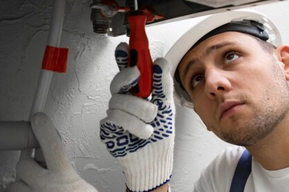 Technik w białym kasku ochronnym i rękawicach roboczych dokonuje naprawy instalacji przy pomocy śrubokręta. Skupiony na pracy w zamkniętym pomieszczeniu.